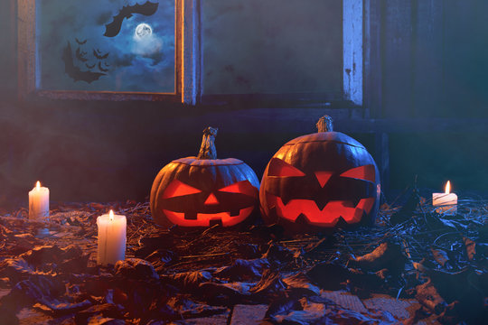Halloween - pumpkins and candles in an abandoned wooden house on leaves and wooden boards with a warm and cold glow, against the background of a window with a mystic sky and bats, in a light smoke