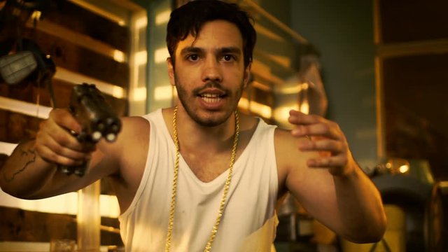 Brutal Gang Member Gone Crazy Makes Threatening Gestures with a Gun. He Wears Gold Chain and Sleeveless Shirt. In the Background Abandoned Building with Boarded Up Windows.