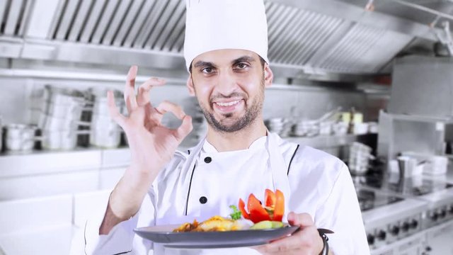 Male Caucasian chef holding and preparing a plate of delicious food while standing and showing OK sign in the kitchen
