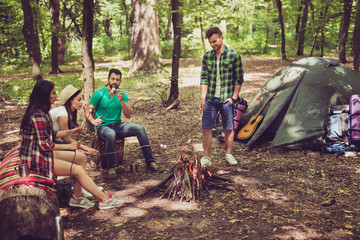Camping, four happy friends are sitting around the fire, roasting the marshmallows, nice sunny day in the forest