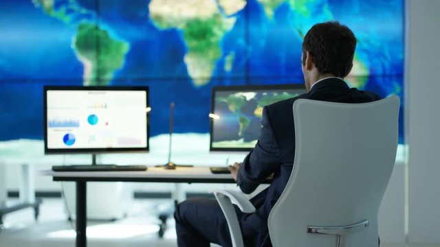 Businessman working in office with large world map graphic on video wall & pie charts & graphs displayed on computer screen. Global business concept.