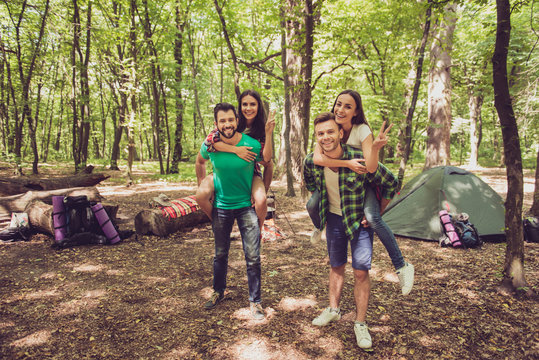 Camp in the sunny wood, tent and backpacks. Two lovely couples, guys are piggybacking their girlfriends, happy, smiling