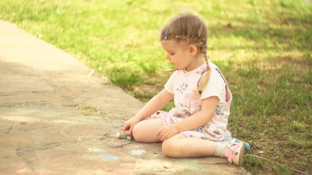 Portrait of a cute little girl dressed in pink overalls, the child draws colorful chalk on the path next to the green grass on a summer sunny day.