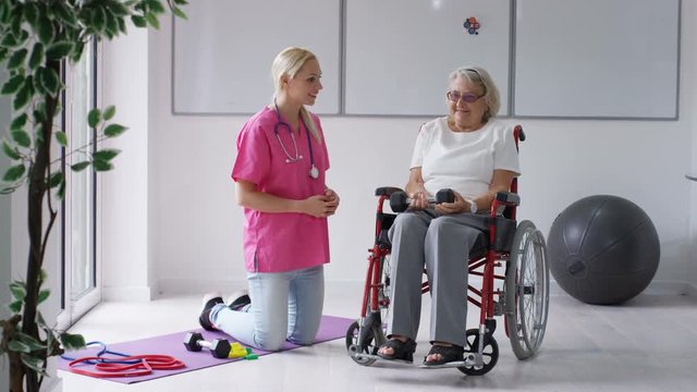 Nurse in residential care home showing elderly lady in wheelchair some exercises with weights.