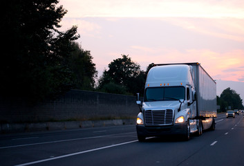 White big rig semi truck with dry van trailer driving on evening road with headlight and sunset sky