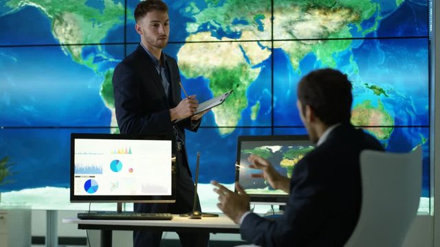  Businessmen discussing large world map graphic on video wall, with pie charts & graphs displayed on computer screen. Global business concept.