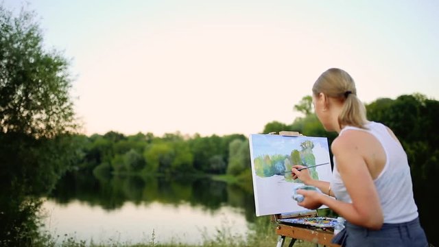 young artist painting picture in park with brushes and colors sitting by the romantic lake and urban buildings in background. ultra wide angle view