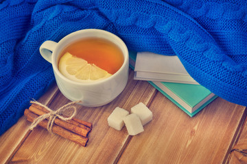 White cup with hot tea and lemon, books and knitted scarf or sweater on wooden table.