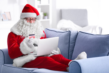 Santa Claus with laptop and credit card on sofa at home