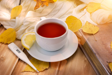 Thermometer and tea with lemon in white cup and old book on a wooden table with autumn yellow leaves