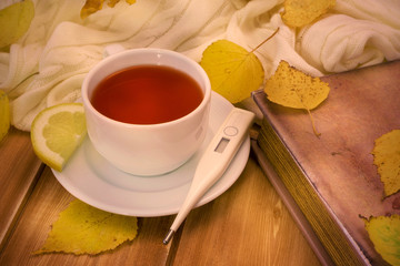 Thermometer and tea with lemon in white cup and old book on a wooden table with autumn yellow leaves. Flu, cold concept