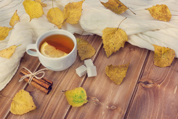 Tea with lemon in white cup, cinnamon sticks, sugar and autumn yellow leaves on wooden background
