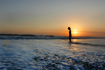 young beautiful Asian girl alone at sea shore looking at orange sky sunset over ocean