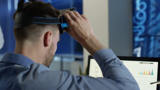 Businessman interacting with a virtual reality headset in futuristic office, computer screens showing financial information & digital code