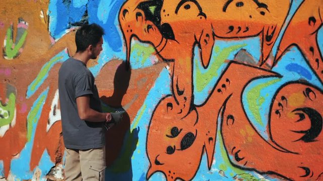 Graffiti Artist Painting With Aerosol. Man with spray bottle. Slow Motion. Young urban painter drawing colorful graffiti on the urban street wall at summer sunny day.
