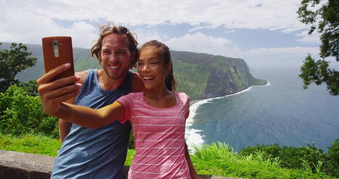 Happy young couple in love having fun taking selfie with phone outside during travel vacation summer holidays. Interracial Asian Caucasian couple laughing and smiling. RED EPIC SLOW MOTION.