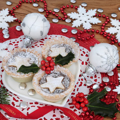 Traditional christmas mince pie tarts on a heart shaped plate with white snowflake and round bauble decorations and holly.