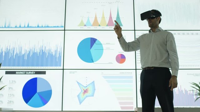  Businessman interacting with a virtual reality headset in front of video wall with financial graphs