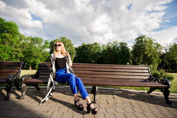 Красивая, стильная девушка со светлыми волосами сидит на скамейке в парке в черной майке и синих брюках 