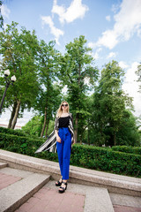 Fototapeta na wymiar Красивая, стильная девушка стоит в парке в черной майке и синих брюках. Девушка со светлыми волосами в солнечных очках 