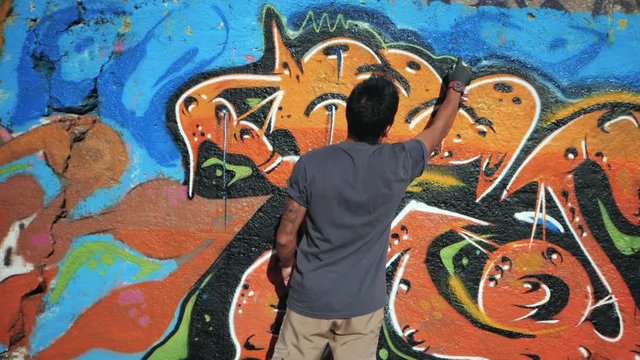 Handsome Talented Young Boy making a colorful graffiti with aerosol spray on urban street wall. Cinematic toned slow motion footage. Creative illegal art. Back view, overall plan