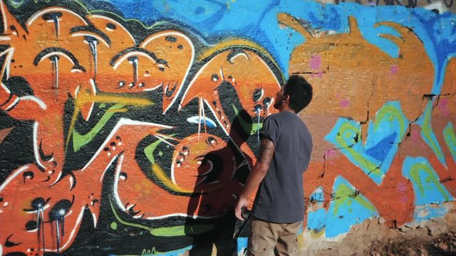Graffiti Artist Painting On The Street Wall. Handsome Man with aerosol spray bottle spraying with colorful paint, Urban Outdoors Art Concept. Slow motion. Gimbal shot. Back view, overall plan