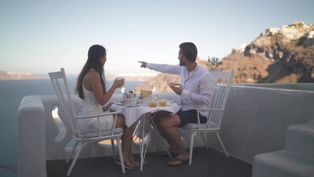 4k travel video couple sitting outside on veranda table enjoying breakfast high up on santorini island with amazing view over the caldera
