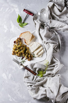 White plate with sliced camembert cheese and baked bunch of green grapes served with bread, wine glass, corkscrew, green leaves, fork on textile over gray texture background. Top view with space