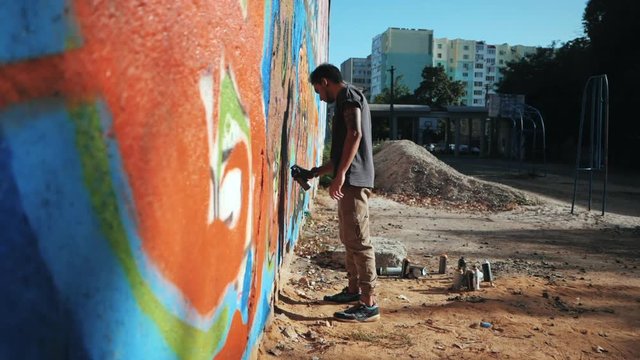 Graffiti Artist Painting With Aerosol. Man with spray bottle. Slow Motion. Young urban painter drawing colorful graffiti on the urban street wall at summer sunny day. Side view