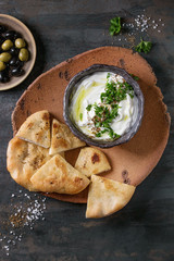 labneh middle eastern lebanese cream cheese dip with olive oil, salt, herbs served with olives, traditional pita bread on terracotta plate over dark texture metal background. Top view with space
