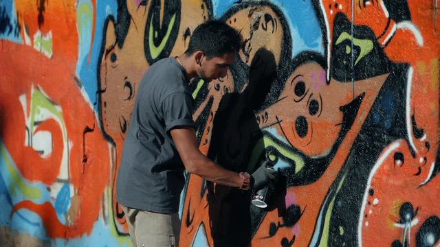 Graffiti Artist Painting On The Street Wall. Handsome Man with aerosol spray bottle spraying with colorful paint, Urban Outdoors Art Concept. Slow motion.