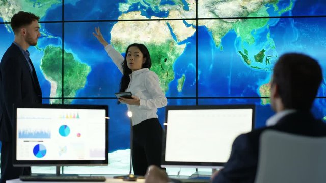  Business team discussing large world map graphic on video wall, with pie charts & graphs displayed on computer screen. Global business concept.
