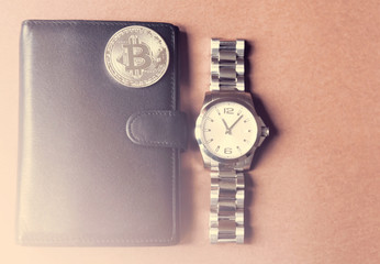 On a brown background is a black wallet, on it is a silver coin bitcoin, beside a man's watch. With a lighting.