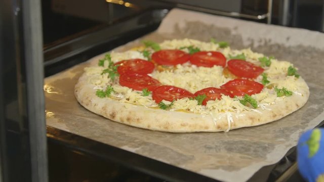 Cooking frozen italian pizza in the oven closeup. Chef puts baking sheet with pizza topped tomatoes mozzarella and edam cheese into the oven. Food preparation in electric cooker.