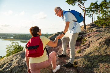 Active senior couple hiking on the top of rock. Mature man helping woman climbing up. Happily smiling. Scenic view of gulf and sea. Healthy lifestyle. Finland.