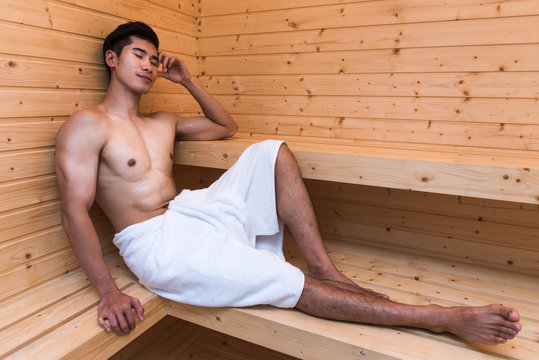 Happy smiling young asian male with a muscular body in towel relaxing in wooden sauna 