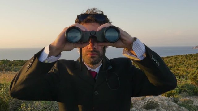 Man in elegant suit with briefcase looking through binoculars on top of a cliff on an island