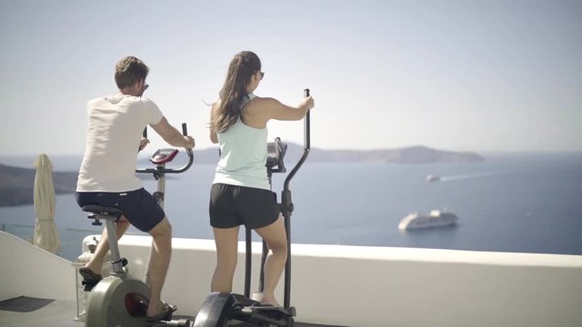 4k travel video happy couple on vacation in santorini on fitness stepper and bike on terrace of luxury hotel facing the caldera
