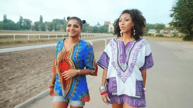 Two happy beautiful afro-american women friends walking outdoor. Young mixed race girls wearing colorful clothing communicates, enjoys the meeting and laughing in slow motion. Dolly shot.