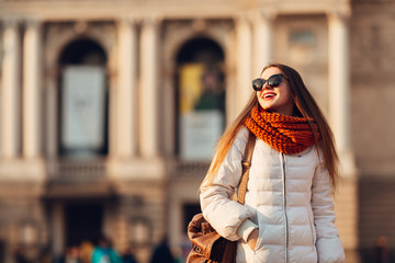Smiling girl in sunglasses wears white down jacket and a orange scarf on the background of old blurred building. Tourism, happiness, trip concept