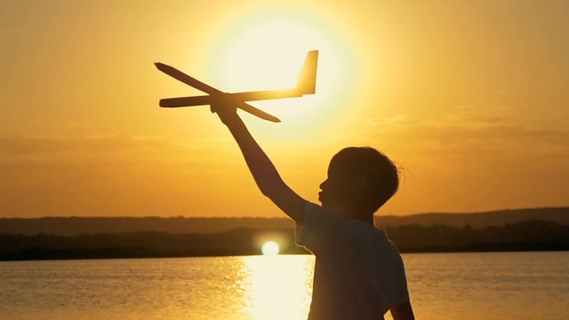 Happy child on a background of an orange sky and lake in summer at sunset, playing with a toy airplane.