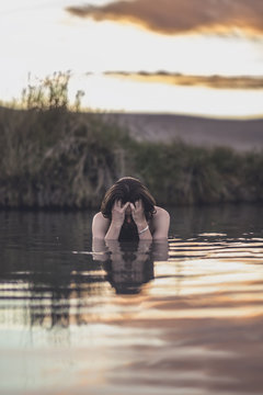 A man in water with long hair covers his face at sunset