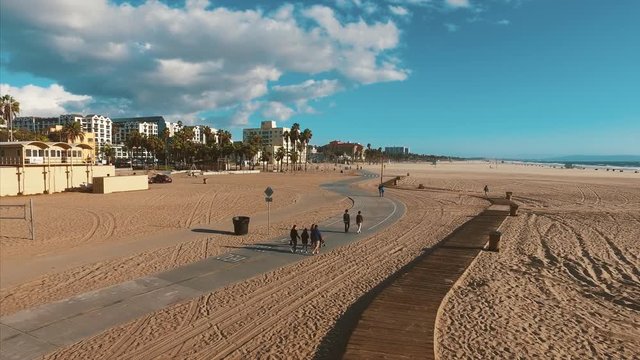 Tourists are walking along the path on the beach near Santa Monica pier. Slow motion. Beautiful sunset. Clouds float on the blue sky.
