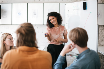 Beautiful African American lady with dark curly hair standing near board and happily looking at her colleagues in office. Young smiling business woman giving presentation to coworkers