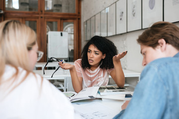 Portrait of upset African American girl with dark curly hair sitting in library with books on table and emotionally discussing something with friends . Group of young tired students studying together