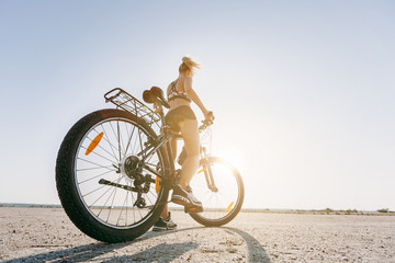 A strong blonde woman in a multicolored suit sits on a bicycle in a desert area and looks at the sun. Fitness concept. Back view. Close-up