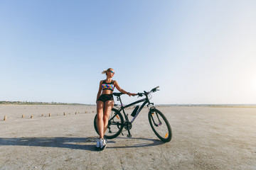 A strong blonde woman in a multicolored suit and sunglasses stands near a bicycle in a desert area and looks at the sun. Fitness concept. Blue sky background