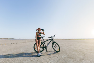 A strong blonde woman in a multicolored suit and sunglasses stands near a bicycle, drinks water from a bottle in a desert area and looks at the sun. Fitness concept. Blue sky background
