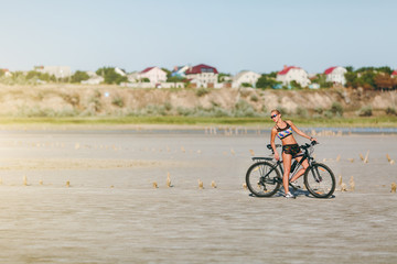 Obraz premium A strong blonde woman in a multicolored suit and sunglasses sits on a bicycle in a desert area. Fitness concept. Blue sky background
