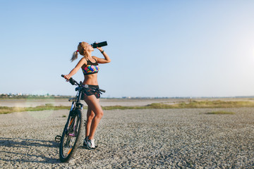 A strong blonde woman in a colorful suit and sunglasses stands near a bicycle, drinks water from a black bottle in a desert area. Fitness concept. Blue sky background